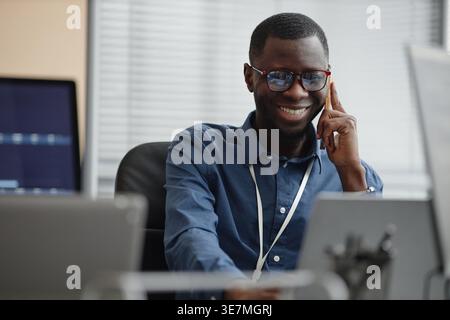 Black male IT specialist wearing eyeglasses having phone call using laptop sitting at desk in office Stock Photo