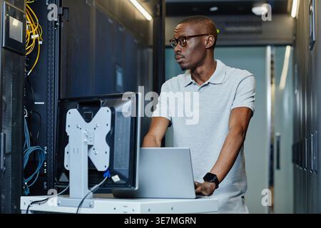 Black male IT engineer with eyeglasses looking at cables standing at table with laptop in server room in data center Stock Photo