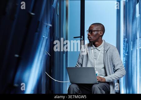 Black male IT engineer with eyeglasses looking at cables using laptop sitting on chair in server room in data center Stock Photo