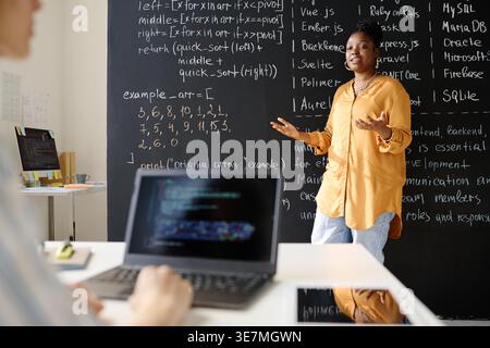Black female IT teacher discussing lesson to students while standing against chalkboard in the classroom Stock Photo