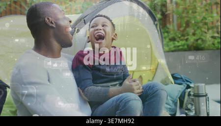 African American dad and son laughing while sitting on lawn at yellow tent, thermos, overlay icons Stock Photo