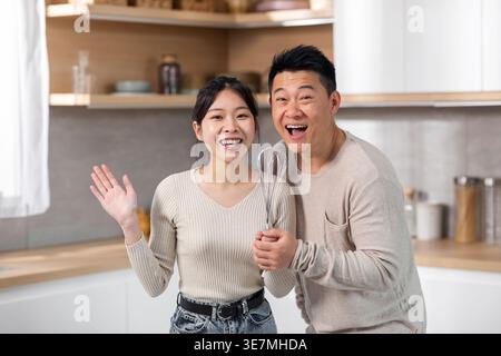Cheerful Asian couple having fun singing using whisk at microphone while standing together in kitchen at home Stock Photo