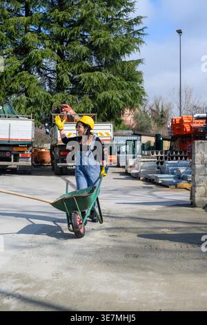 Happy Hispanic female worker wearing denim overalls, yellow hardhat, and protective gloves pushing wheelbarrow with shovel with one hand at constru... Stock Photo