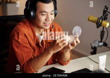 Happy Asian male online seller wearing headset and holding foundation sitting at table with makeup and skincare products streaming live from home s... Stock Photo