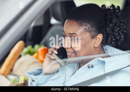 Black woman wearing seatbelt talking on smartphone going home after grocery shopping sitting on driver's seat in car during daytime Stock Photo