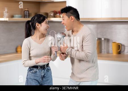 Cheerful Asian couple having fun singing using kitchen utensils as microphones while standing together at home Stock Photo