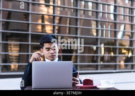 Serious Asian businessman smoking cigar and using smLifestylephone sitting at table with laptop, coffee, and slice of cake on plate in outdoor cafe durin... Stock Photo