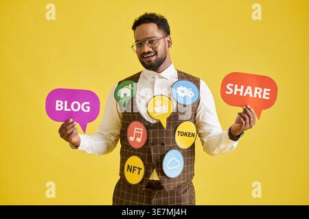 Smiling Asian male influencer showing paper icons standing against yellow background in studio Stock Photo