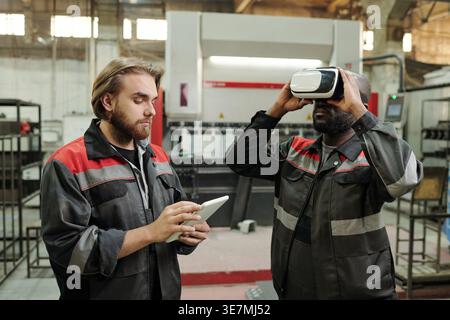 Black male engineer wearing VR headset standing near caucasian male colleague standing inside factory against hydraulic press brake machine in the ... Stock Photo