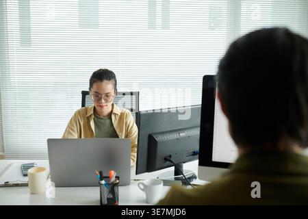 Asian female programmer wearing eyeglasses working on laptop sitting at table near male colleague in office Stock Photo
