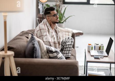 Sick Arab man wrapped in blanket holding neck sitting on sofa near table with laptop and medicine at home Stock Photo