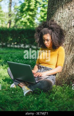 Hispanic female with curly hair working on laptop sitting on grass leaning on tree trunk in park during daytime Stock Photo