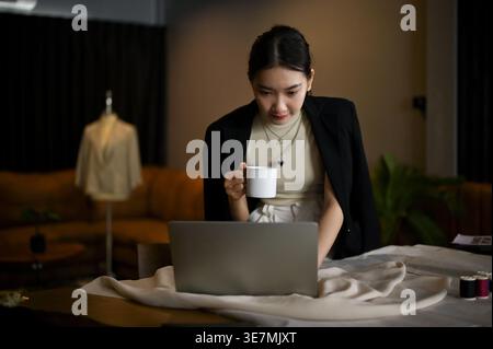 Asian female fashion designer having coffee while using laptop in tailor shop Stock Photo