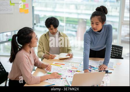 Asian female UX/UI designer leaning on table using laptop working with teammates in office Stock Photo