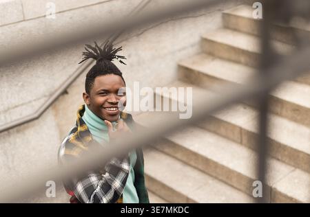 Smiling Black man with locks wearing turtleneck sweater and checkered shirt touching chin while standing on stairs in city Stock Photo