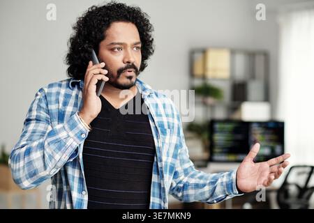 Serious Indian man having phone call standing near computer in office Stock Photo