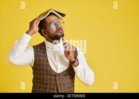 Confused Asian man wearing eyeglasses covering head with book looking up pointing pen on chin standing against yellow background in studio Stock Photo