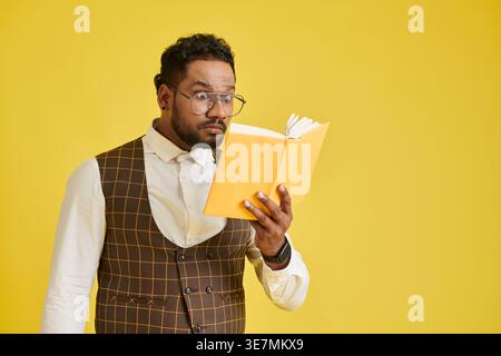 Shocked Asian man wearing eyeglasses reading book standing against yellow background in studio Stock Photo