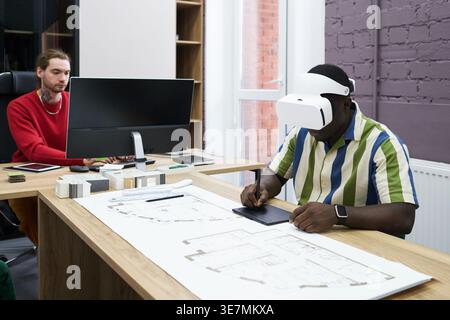 Black man wearing virtual reality headset using graphic tablet sitting at table with mixed race coworker using computer in office Stock Photo