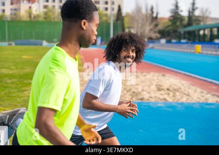 Black male runners stretching legs on barrier standing on running track on sunny day Stock Photo