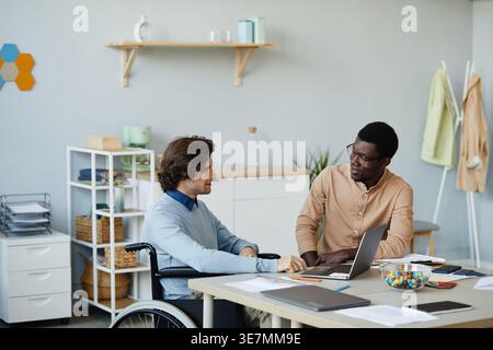 Black male IT specialist having conversation with disabled male colleague while sitting at table in office Stock Photo