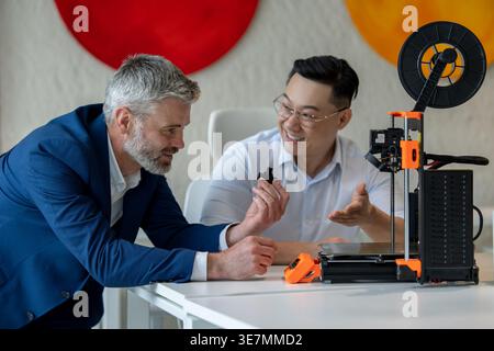 Asian and caucasian male industrial engineers examining product created by 3D printer while sitting at table working together in office Stock Photo