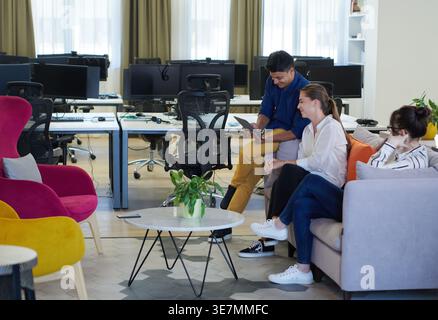 Middle Eastern man using tablet sitting on sofa beside caucasian female coworkers talking in office during break Stock Photo