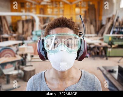 Black female carpenter wearing safety glasses and mask standing in woodworking shop Stock Photo