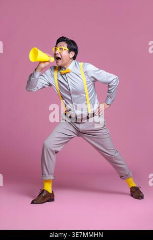 Chinese senior man with eyeglasses looking away standing with legs wide open and hand on waist while shouting through megaphone against pink backgr... Stock Photo