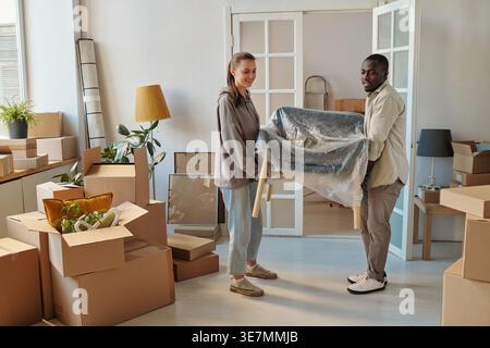 Black man standing and carrying new armchair with caucasian girlfriend during relocation in their new home Stock Photo