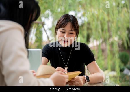 Asian woman working remotely with her colleague while sitting at table in garden during daytime Stock Photo