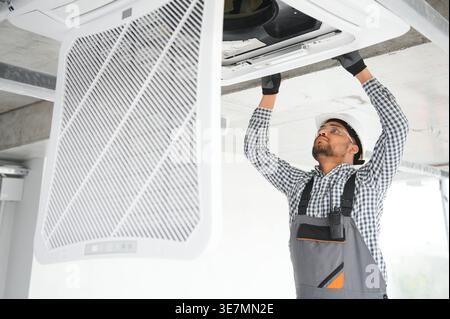 Indian male technician wearing uniform and hard hat checking air conditioner inside the building Stock Photo