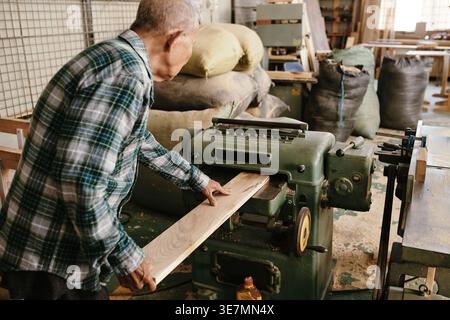 Senior Asian male carpenter wearing checkered long sleeves using thickness planer machine to trim board while standing in workshop Stock Photo