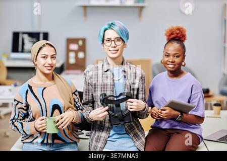 Group of programmers holding virtual headset, and tablet while sitting together on table in office Stock Photo