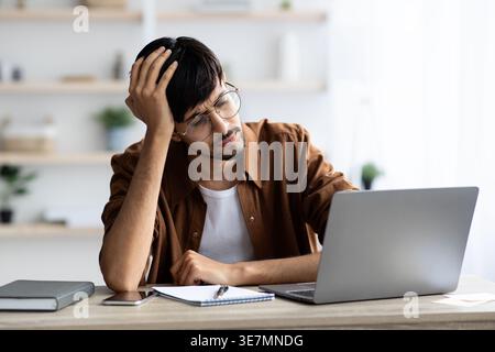 Exhausted indian man sitting in front of laptop at office, touching his head, suffering from burnout or financial crisis, young guy freelancer having Stock Photo