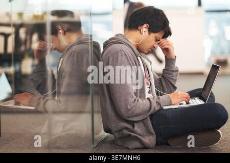 Stressed mixed-race male employee wearing earphones touching face using laptop leaning against glass wall sitting with closed eyes and crossed legs... Stock Photo
