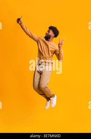 Black man with afro using smartphone taking selfie doing peace sign gesture jumping against yellow background in studio Stock Photo