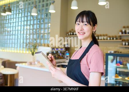 Asian waitress in black apron standing with notepad taking orders from customers inside coffee shop Stock Photo