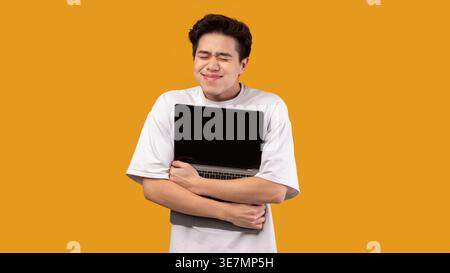 Smiling Asian man wearing white shirt hugging his laptop while standing with closed eyes against orange wall in studio Stock Photo