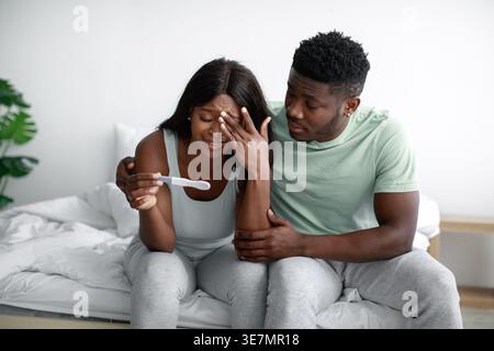 Depressed Black couple holding and looking pregnancy test kit while sitting on bed in bedroom at home Stock Photo