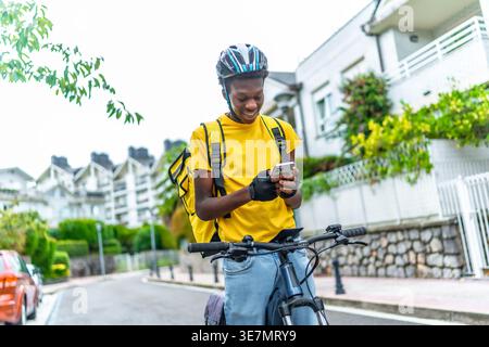 Black man wearing helmet and delivery bag using smartphone to contact customer while standing with bicycle on street during daytime Stock Photo