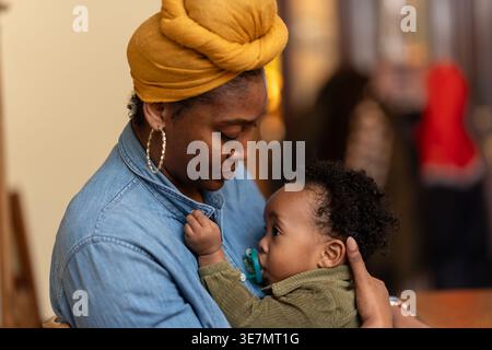 Black mother wearing yellow head wrap and long sleeve denim shirt holding and playing with her baby boy with pacifier while sitting at home Stock Photo