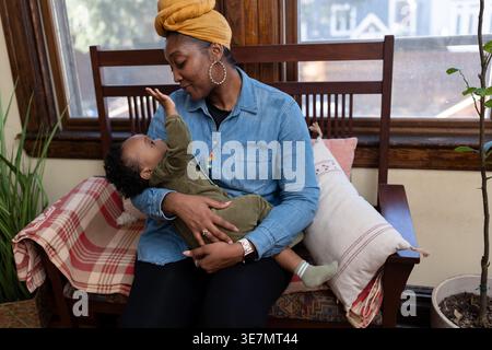 Black mother wearing yellow head wrap and long sleeve denim shirt carrying and playing with her baby boy with pacifier while sitting on wooden sofa... Stock Photo