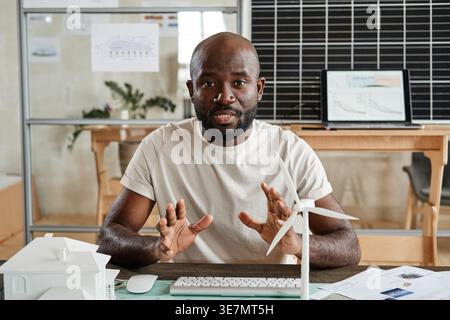 Black male architect with beard wearing white shirt sitting at table gesturing while talking during online meeting in office Stock Photo