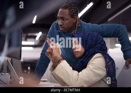 Black male employee standing beside female colleague leaning arms on table discussing and using desktop sitting at table in office Stock Photo