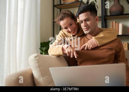 Smiling mixed race woman pointing finger and embracing Asian boyfriend from behind using laptop sitting on sofa in living room at home Stock Photo