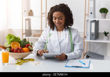 Black female dietitian using tablet while sitting at table with bowl of fruits and vegetables, orange juice, and clipboard in clinic Stock Photo