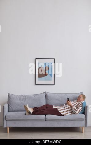 Vertical wide angle portrait at modern mixed-race woman using smartphone while lying on sofa in minimal home interior, copy space Stock Photo
