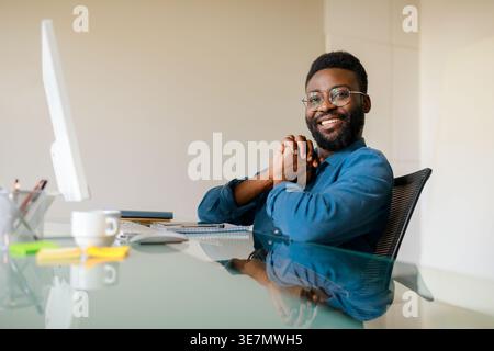 Black bearded man wearing eyeglasses with clasped hands sitting at table in office Stock Photo