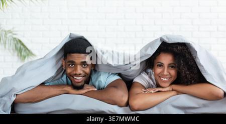Smiling Black couple leaning chins on their hands while lying together on bed under blanket at home Stock Photo